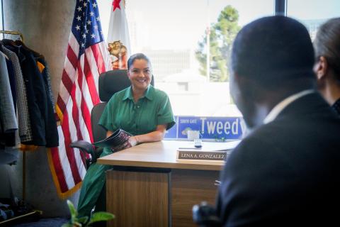 Smiling woman in green dress sits at office desk with two flags behind her.